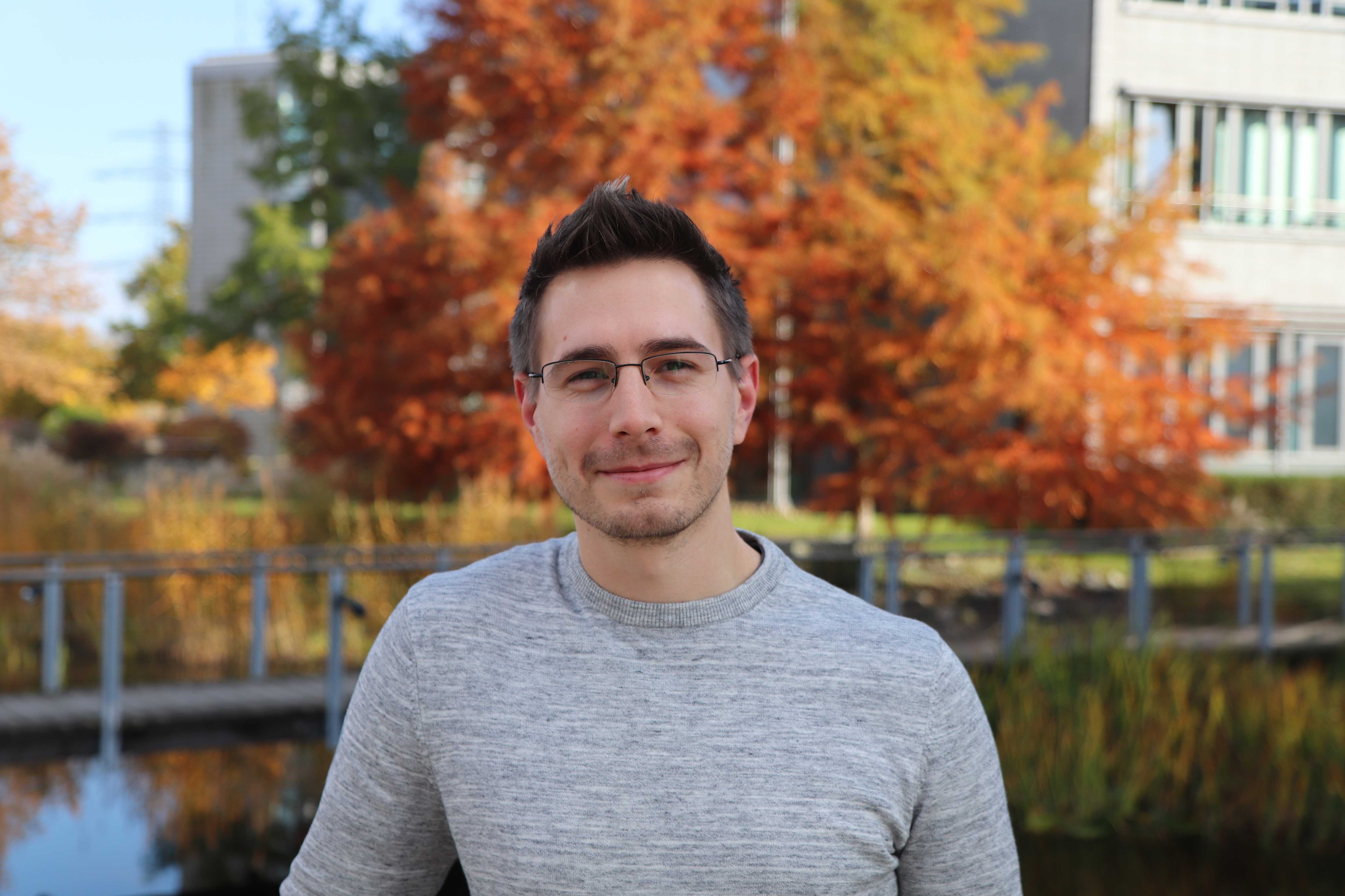 Ein junger Mann mit Brille, grau-meliertem Pullover und dunklen Haaren lächelt in der Herbstsonne in die Kamera. Hinter ihm befindet sich ein Baum mit Herbstlaub vor einem grauen Bürogebäude.