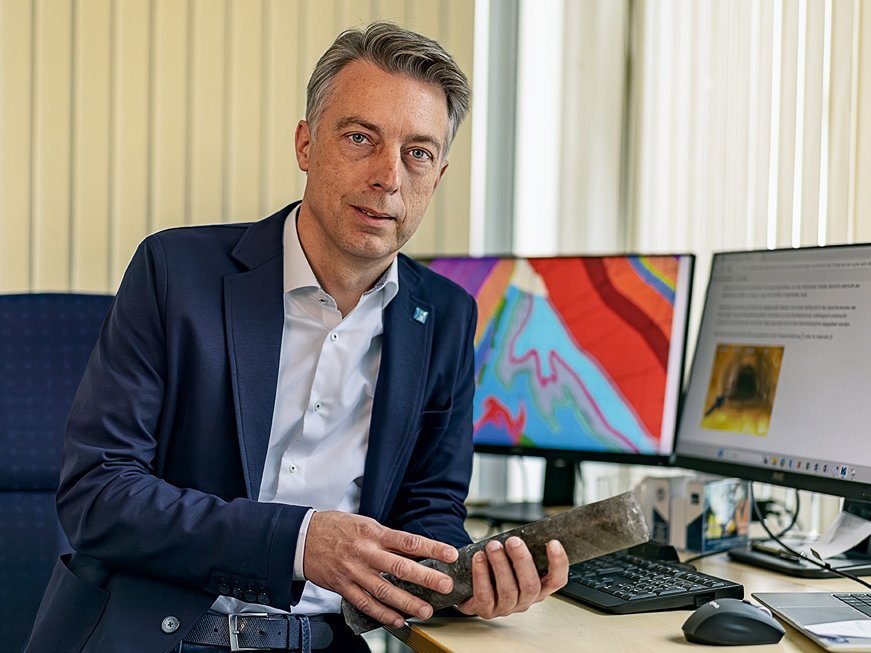 An einem Schreibtisch mit zwei Bildschirmen sitzt ein Mann mit kurzen grauen Haaren. Er trägt ein weißes Hemd unter einem dunkelblauen Sakko und lehnt mit einem Ellbogen auf dem Tisch. 