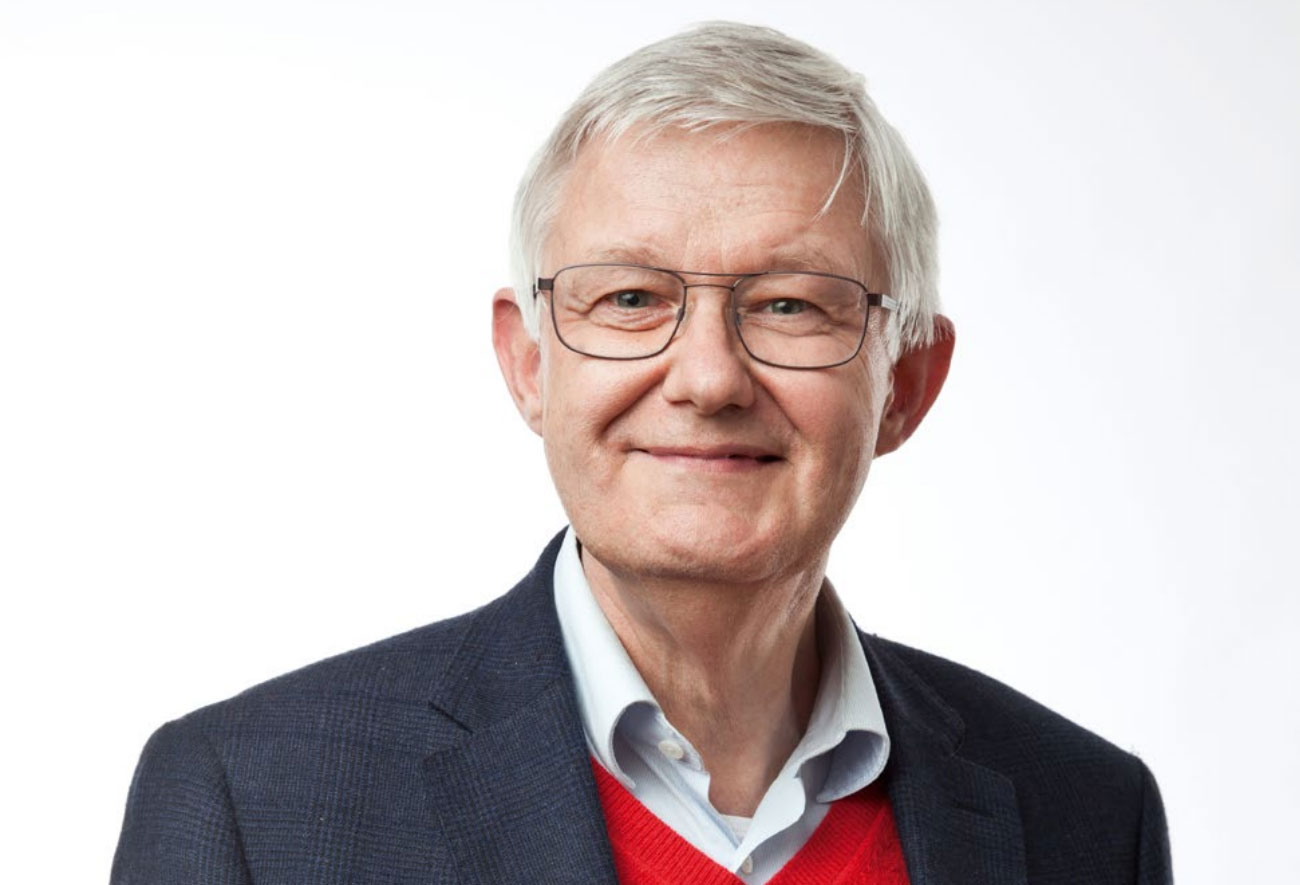 Portraitfoto eines Mannes mit kurzen grauen Haaren und Brille. Er trägt ein dunkelgraues Sakko und darunter ein Hemd sowie einen roten Pullover mit V-Ausschnitt.