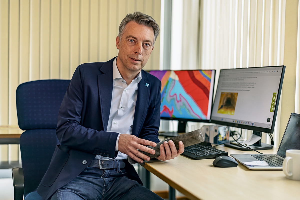 An einem Schreibtisch mit zwei Bildschirmen sitzt ein Mann mit kurzen grauen Haaren. Er trägt ein weißes Hemd unter einem dunkelblauen Sakko und lehnt mit einem Ellbogen auf dem Tisch. 
