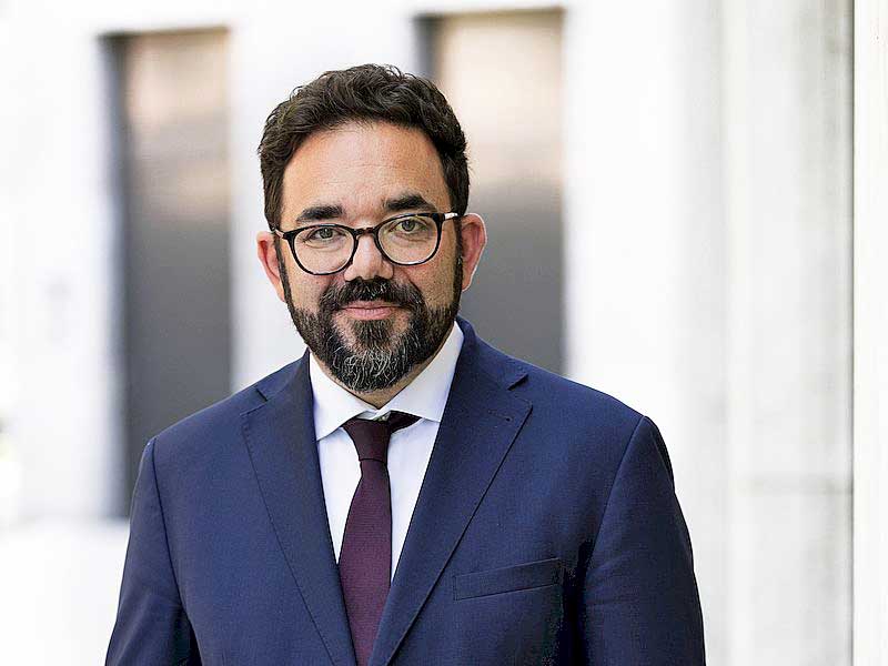 Ein Mann mit schwarzen Haaren, Bart und Brille blickt frontal in die Kamera. Er trägt einen dunkelblauen Anzug und dazu ein weißes Hemd sowie eine Krawatte.