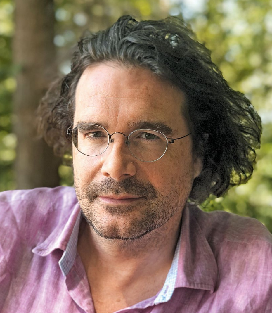 Portraitfoto eines Mannes mit dunklen, halblangen und nach hinten gekämmten Locken. Er trägt eine Brille mit runden Gläsern und ein rosa kariertes Hemd sowie einen Dreitagebart.